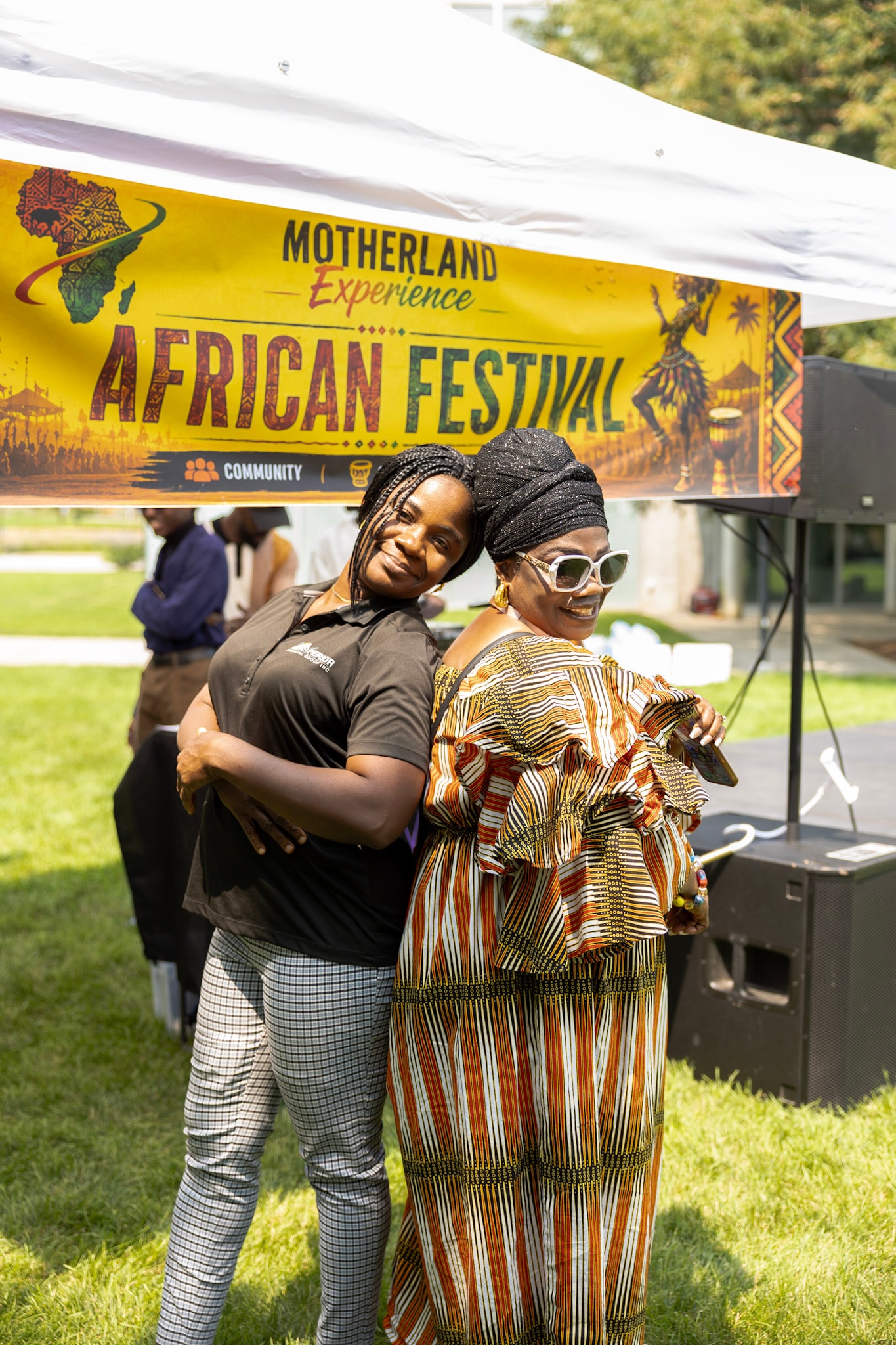 Two women posing back-to-back in front of the Motherland Experience African Festival banner