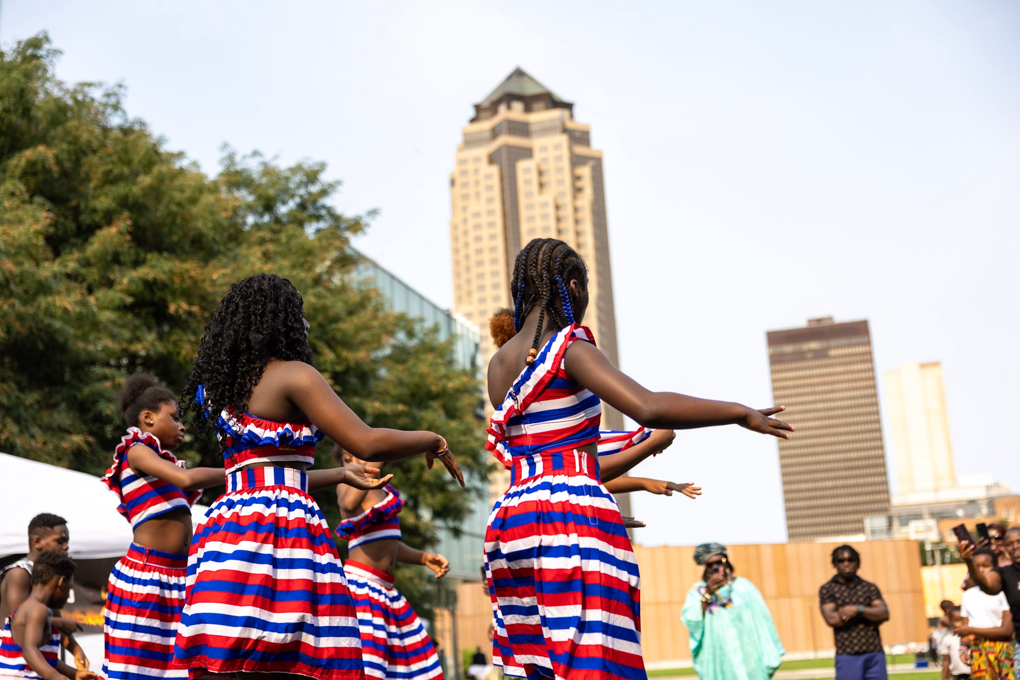 Young dancers in red, white and blue striped outfits performing with the Des Moines skyline behind them