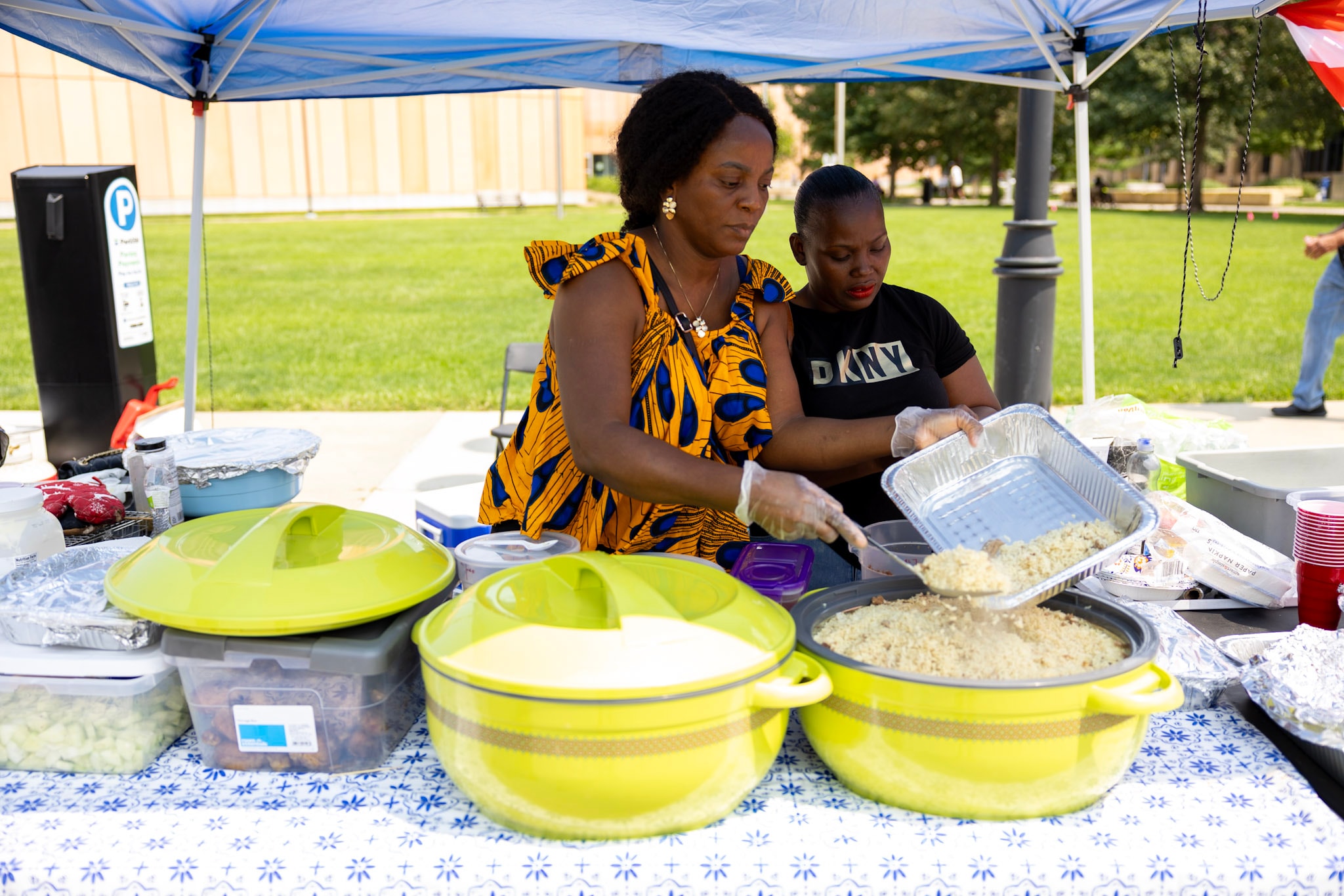 Two women in African print attire serving food from large bowls at a vendor tent